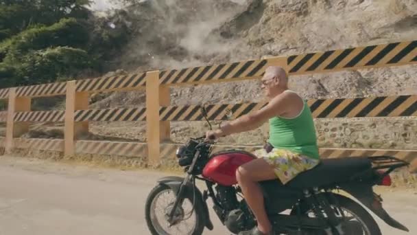 Старший едет на мотоцикле мимо гор с паром из горячих источников. Взрослый человек путешествует на мотоцикле по горному ландшафту с термальными источниками . — стоковое видео