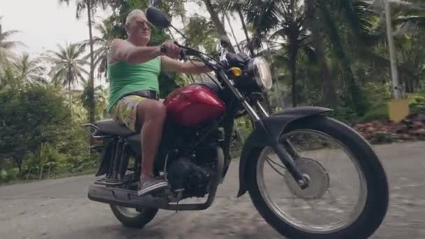 Pensioner riding motorcycle on tropical village at sunny day. Elderly man motorbiker driving on motorcycle on tropical nature background. Moto travel concept. — Stock Video