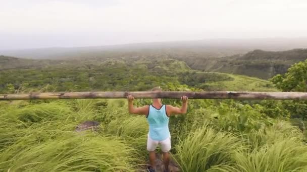 Людський пауерліфтер використовує дерев'яний брусок для тренувань у тропічному ландшафті. Спортсмен піднімає дерев'яний штангу для тренувань з бодібілдингу на зеленому пагорбі та на високогір'ї. Зовнішній ваговий підйом . — стокове відео