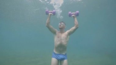 Young athletic sportsman in mask is making shoulders exercises with dumbbells underwater, front view.