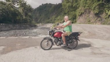 Yaz tatilinde moto gezisi sırasında kırsal yolda motosiklet üzerinde sürüş yaşlı adam. Asya köyünde nehir köprüsünde motosiklete binen gezgin adam. Moto seyahat.