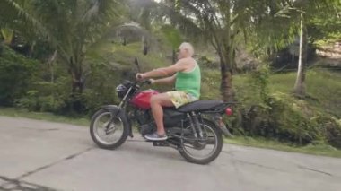 Yetişkin adam yeşil dağlık arazide tropikal yolda motosiklet sürme. Yaz tatilinde moto seyahat ederken motosiklete binen emekli adam. Motosiklet seyahat.