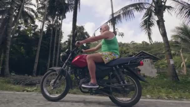 Reifer Mann auf Motorrad unterwegs auf Dorfstraße auf tropischen Palmen Landschaft. Senior fährt in den Sommerferien Motorrad auf Landstraße. — Stockvideo