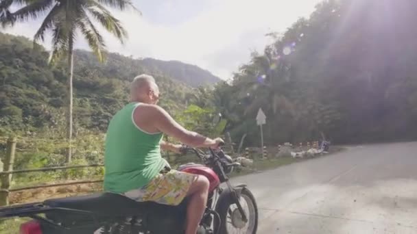 Pria dewasa mengendarai sepeda motor di jalan pedesaan di bukit hijau menutupi hutan tropis. Traveling pria naik sepeda motor di jalan pinggiran kota. Konsep senior aktif . — Stok Video