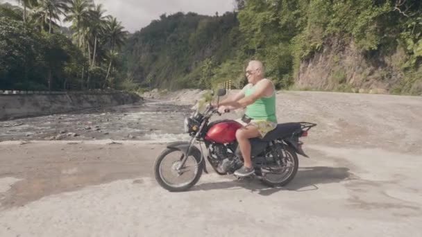 Ηλικιωμένος άνθρωπος οδήγηση με μοτοσικλέτα στο δρόμο της υπαίθρου, ενώ το ταξίδι με Moto στις καλοκαιρινές διακοπές. Ταξιδευτής με μοτοσικλέτα στη γέφυρα του ποταμού στο ασιατικό χωριό. Ταξίδια με Moto. — Αρχείο Βίντεο