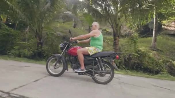 成年男子骑摩托车在热带道路上的绿色高地景观。养老金领取者骑摩托车，而摩托车旅行在夏季。摩托车旅行. — 图库视频影像