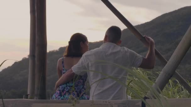 Молодой человек обнимает женщину, сидя в беседке на зеленом горном ландшафте. Любовники купе наслаждаются романтическим свиданием на тропическом холме и горном фоне. Романтические отношения . — стоковое видео