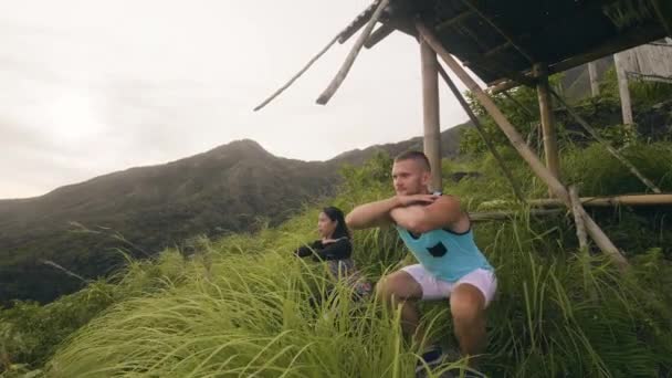 Atleta hombre y la mujer en forma ejercicio sentadilla entrenamiento sobre fondo de montaña tropical. Pareja deportiva en cuclillas mientras entrenan juntos en el paisaje tropical. Entrenamiento al aire libre . — Vídeos de Stock