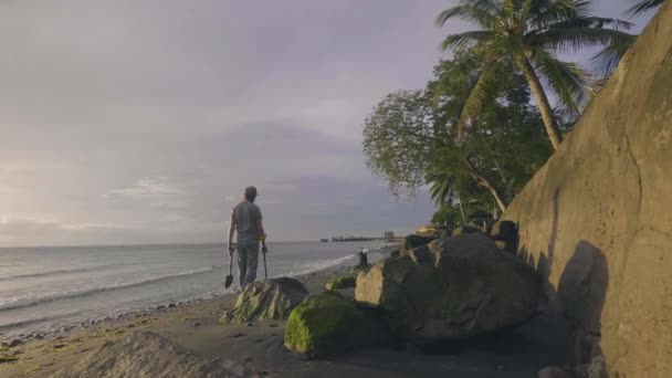 El hombre está escaneando la playa de arena por detector de metales y paseando con su perro al atardecer cerca del océano . — Vídeo de stock