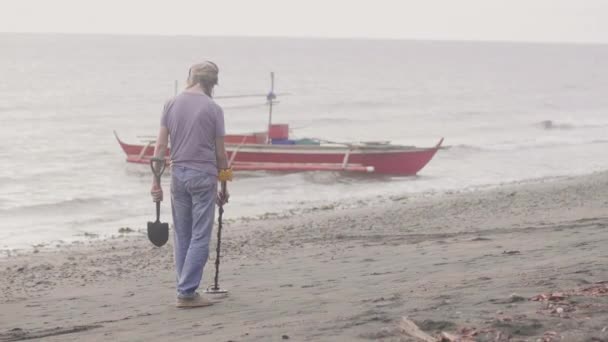 이어폰을 입은 남자가 해돋이에 해안선의 모래에서 귀중품을 찾는 탐지기를 들고 해변에서 일하고 있습니다.. — 비디오