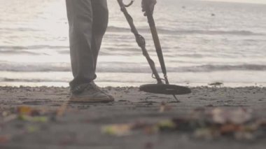 Adam sahilde kumları metal dedektörüyle tarıyor ve sahil şeridinde kazmaya başlıyor..