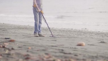 Adam gün doğumunda metal dedektörü kullanarak deniz plajındaki kumda değerli eşyalar buluyor, bacaklar yakın çekimde.