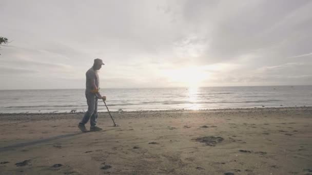 Człowiek w słuchawkach działa skanowanie piasku na plaży z wykrywaczem metali znalezienie kosztowności na wschód słońca. — Wideo stockowe