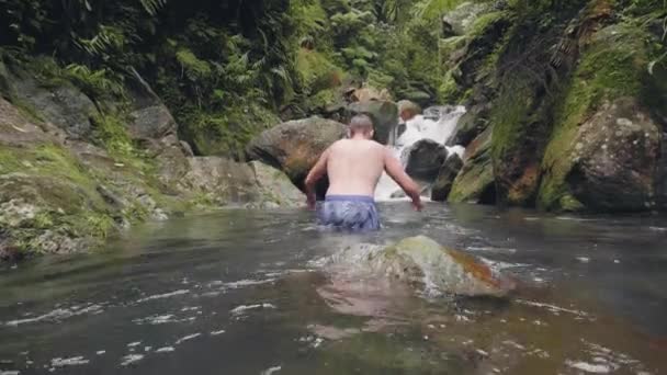 Ung man simmar på flodvatten som rinner från tropiskt vattenfall i regnskogen. Glad man njuter av vattendrag i vattenfall flod i tropisk skog i djungeln. — Stockvideo