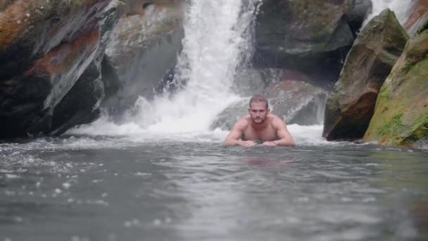 Hombre guapo disfrutando de nadar en el río de montaña desde la cascada tropical en la selva tropical. Hombre joven bañándose en el río rocoso en el paisaje de cascada de montaña en la selva salvaje . — Vídeo de stock