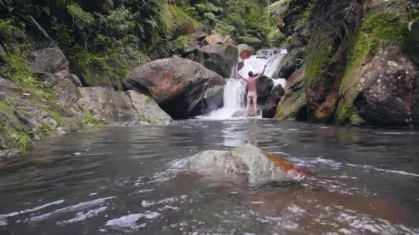 Голий чоловік стоїть з піднятими руками на скелястому водоспаді в тропічних тропічних лісах. Щасливий чоловік розтягує руки над головою насолоджуючись прісною водоспадом у диких джунглях . — стокове відео