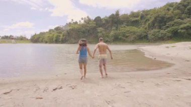 Mutlu çift el ele tutuşuyor ve yaz kamp sırasında göl suyunda koşuyor. Yürüyüş tatili sırasında yaz gölünde su sıçrayan neşeli erkek ve kadın.