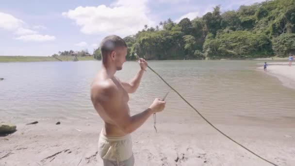 Hombre joven utilizando cuchillo de supervivencia y palo de la planta para hacer caña de pescar en la orilla del lago. Joven cazador con cuchillo haciendo flecha desde palo de madera para cazar. Concepto de pesca y caza . — Vídeos de Stock