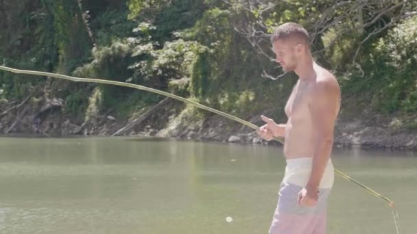 Fisher hombre con caña de pescar de madera en el lago tropical y cascada en el fondo. Joven pescando pescado con carretera de pesca hecha a mano en la orilla del lago vacía . — Vídeos de Stock