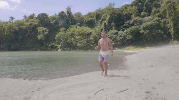 Bonito homem com vara de pesca artesanal andando na costa do lago na paisagem tropical da natureza. Jovem pescador com equipamento de pesca na praia tropical vazia na ilha selvagem . — Vídeo de Stock