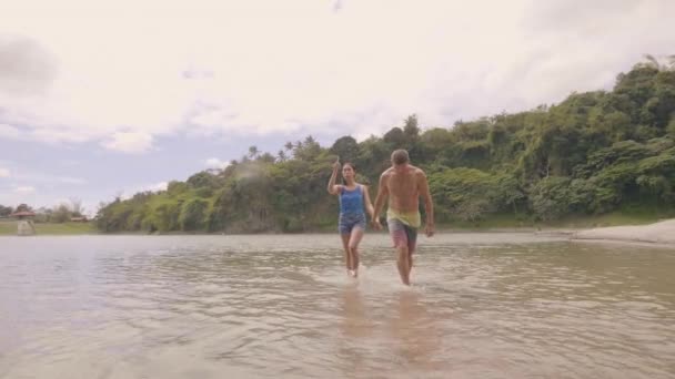 Asyalı kadın ve Avrupalı adam el ele tutuşuyor ve tropikal doğa manzarasında göl suyunda yürüyorlar. Çok ırklı çift göl kıyısında yaz tatili harcama, yürüyüş ve doğa izliyor. — Stok video