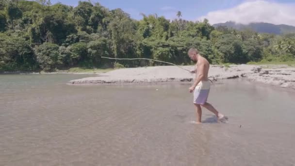 Белый мужчина с удочкой ручной работы ходит по воде и смотрит на рыбу на острове выживания. Молодой человек с деревянными рыболовецкими снастями собирается ловить рыбу в озере . — стоковое видео