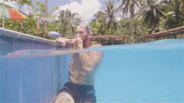 Young swimmer man emerges from outdoor swimming pool in resort hotel waterline view. Bearded man sitting on poolside on summer hotel. Summer activity.