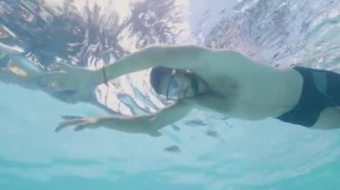Young man in glasses swimming in outdoor pool while summer vacation at resort hotel. Underwater view face bearded man in mask swimming in clear water on tropical palm background