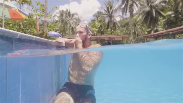 Young swimmer man emerges from outdoor swimming pool in resort hotel waterline view. Bearded man sitting on poolside on summer hotel. Summer activity. — Stockvideo