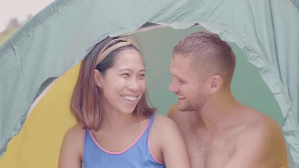 Happy asian woman and european man smiling and talking in camping tent at weekend. Traveling couple sitting inside tourist tent while travel at summer hike. Tourism and travel concept. — Stock Video
