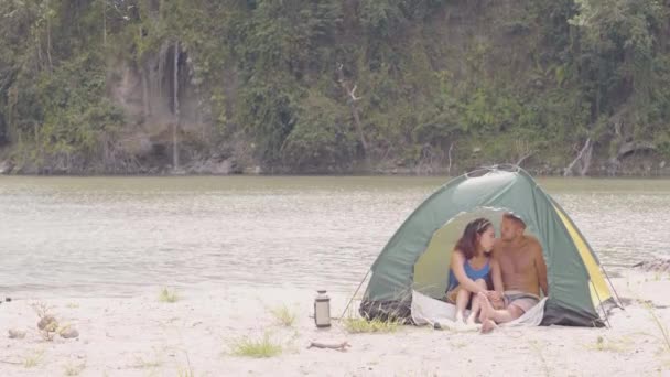 Romantiskt par i kärlek sitter i turist tält på sjön stranden och berget vattenfall landskap. Ungt turist par kysser och omfamnar i camping tält medan sommar vandring till berget. — Stockvideo