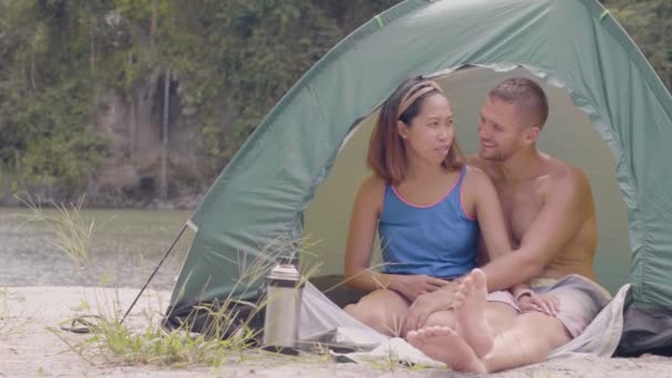 Young couple in love resting in tourist tent on tropical waterfall and lake landscape. Smiling asian woman and caucasian man sitting in camping tent at lake shore while summer hike. — 비디오