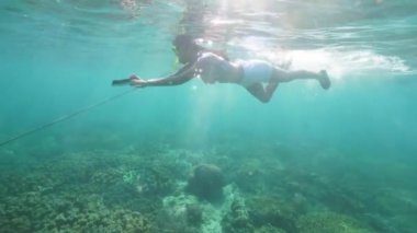 Şeffaf deniz suyunda şnorkel maskesi ve mobil kamerasıyla şnorkelle yüzen genç bir kadın. Mavi denizde şnorkel maskesi ve tüple yüzen su altı kızı..
