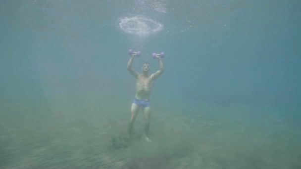 Athletic man does sport exercise with dumbbells underwater stands on ocean floor. — Stock Video