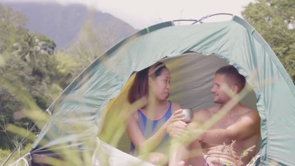 Feliz casal de raças mistas conversando e bebendo chá na tenda turística enquanto viaja de verão. Casal de turistas sorridentes conversando e bebendo chá dentro da barraca de acampamento na paisagem verde da montanha e da floresta . — Vídeo de Stock