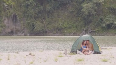 Mutlu karışık ırk çift birlikte yaz tatili harcama. Turist çift göl kıyısında kamp çadırında oturan ve yeşil dağ manzara dan şelale akan. Yaz yürüyüşü, seyahat ve seyahat.