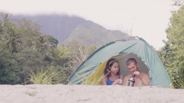 Pareja multinacional bebiendo té caliente del termo en el interior de la tienda de campaña turística en la montaña verde y el paisaje forestal. Caucásico hombre verter té de termo botella a asiático mujer en camping tienda de campaña . — Vídeos de Stock