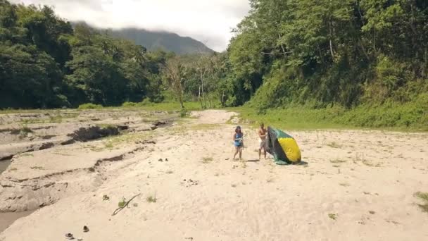 Traveling couple setting tourist tent at campsite while summer hike. Young man and woman setting camping tent on lake shore and rainforest and green mountains background. — Stock Video