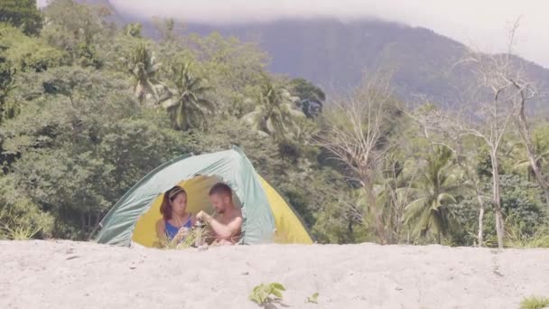 Uomo e donna stanno bevendo un tè dal thermos seduti in tenda sullo sfondo della montagna . — Video Stock