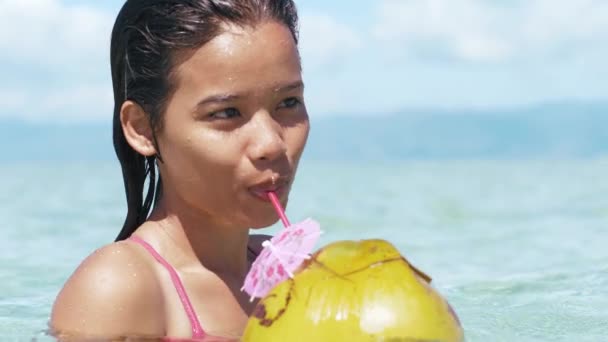 Closeup portrait of pretty asian girl in swimsuit drinks coconut on ocean beach. — Stock Video