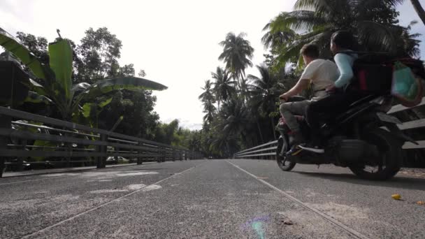 Visão traseira de homem e mulher andando de scooter atravessando uma ponte debaixo de árvores . — Vídeo de Stock