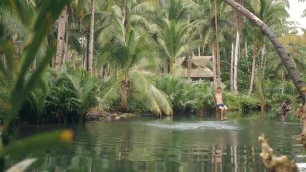 Caucasian man enjoys rope swingong back and forth on coconut tree into a river. — Stock Video
