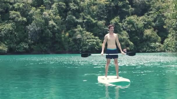 Um homem bonito jovem feliz paddleboarding em uma lagoa cênica e serena na Ásia . — Vídeo de Stock
