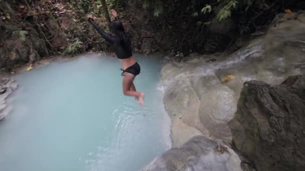 Een vrouw in badpak springt van de rand van de waterval in een turkoois water.. — Stockvideo