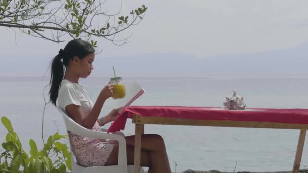 Teenager woman drinking juice while reading book at the table in the beach cafe — Stock Video