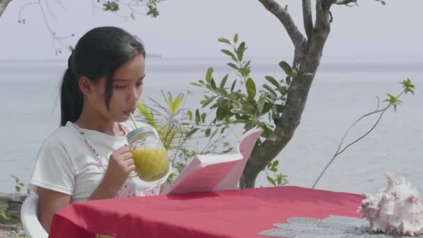 Hermosa joven sentada en una mesa roja bebiendo jugo y leyendo un libro . — Vídeos de Stock
