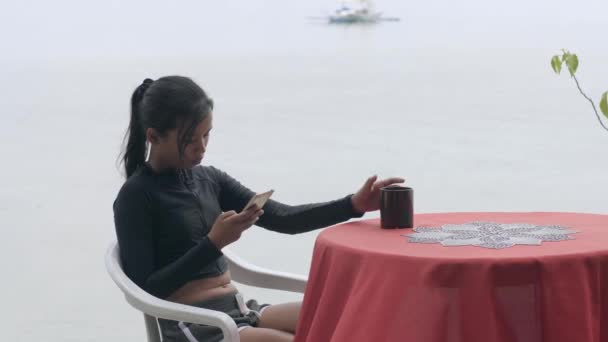 Een meisje zit aan tafel in een café vlakbij het strand met telefoon en koffie. — Stockvideo