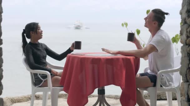 Cheerful young couple sitting at a table near the beach enjoys drinking coffee. — Stock Video