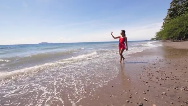 Deniz kıyısında video bloğu çeken kırmızı elbiseli bir kadın gezgin. . — Stok video