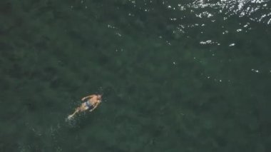 Daire çizen dron görüntüsü: Temiz suda yüzen ve yüzen genç erkeksi bir adam.